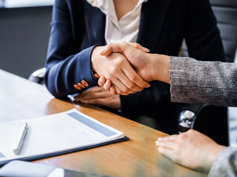Two female lawyers in business suits shake hands at wooden desk, finalizing legal agreement. wooden gavel and scales represent justice as they discuss corporate law, compliance, dispute resolution.